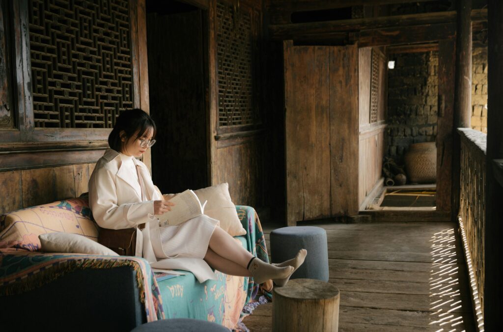 Asian woman sitting on a sofa reading a book on a wooden patio, surrounded by rustic decor and natural light.