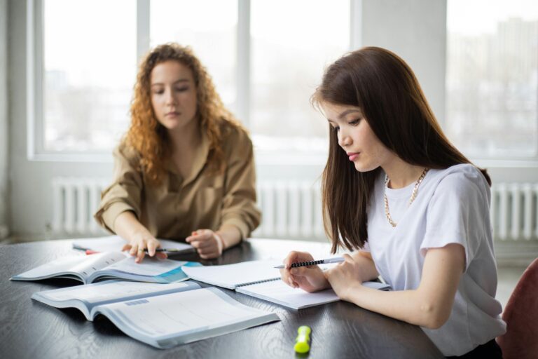 Serious multiracial female students writing in notebooks at table with textbook while preparing for exam together in classroom during lesson